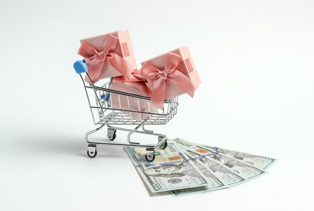 Shopping trolley with gift boxes and one hundred dollar bills on a white background.