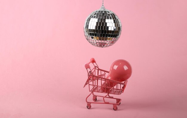 Shopping trolley with light bulb and disco ball on pink background Minimalism party concept