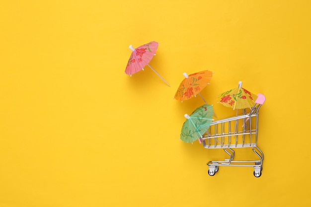 Shopping trolley with Mini cocktail umbrellas on yellow background Summer background Top view