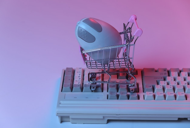 Тележка для покупок с компьютерной мышью на старой клавиатуре. Розовый синий градиент неона, голографический свет. Атрибуты 80-х, ретро волна