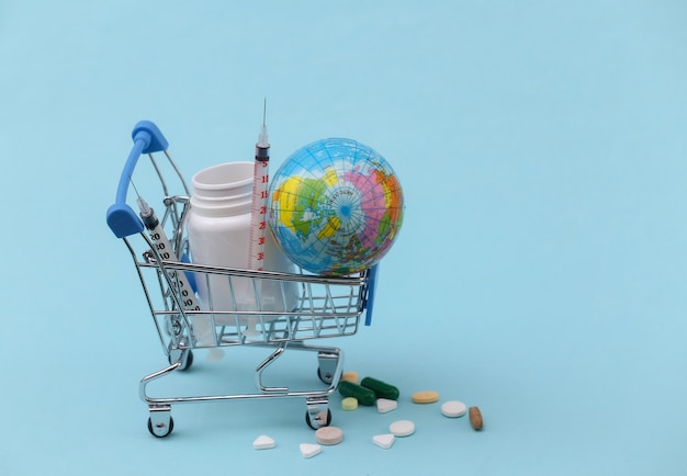 Shopping trolley with pills bottle, syringe and globe on blue background.