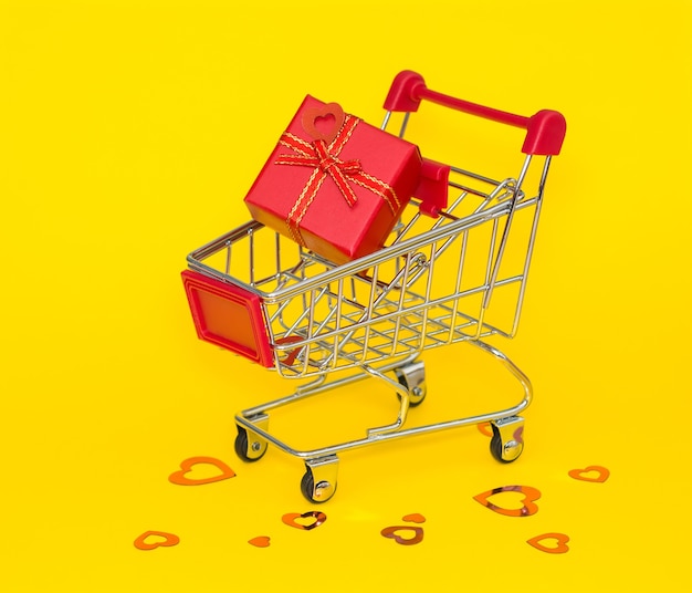 Shopping trolley with red gift and red confetti on a yellow background.