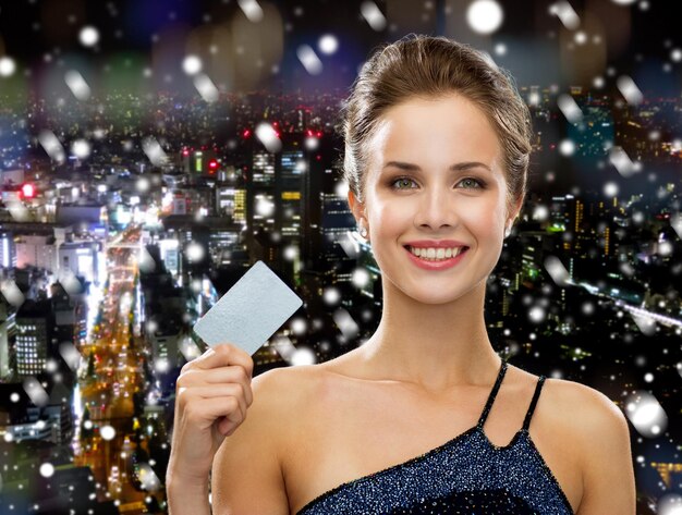 쇼핑, 부, 크리스마스, 휴일 및 사람들 개념 - 눈 덮인 밤 도시 배경 위에 신용 카드를 들고 이브닝 드레스에 웃는 여자
