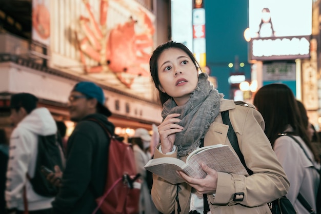 Shopping young girl reading guidebook in modern urban.\
beautiful woman having fun in city in dotonbori. female tourist\
standing in crowded searching direction on map in travel book at\
night city.