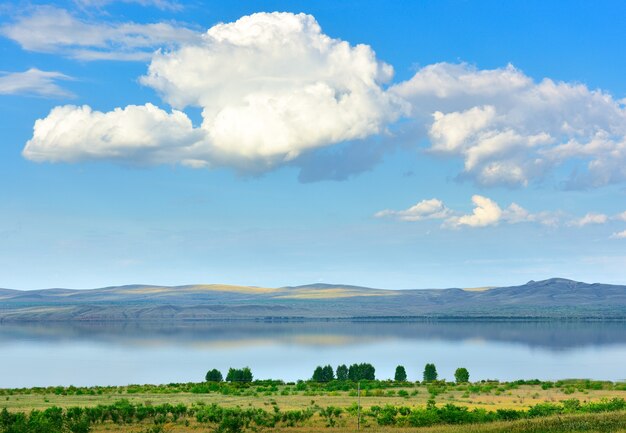 The shore of a steppe reservoir among blue mountains in summer under a big white cloud in a blue sky. Siberia, Russia