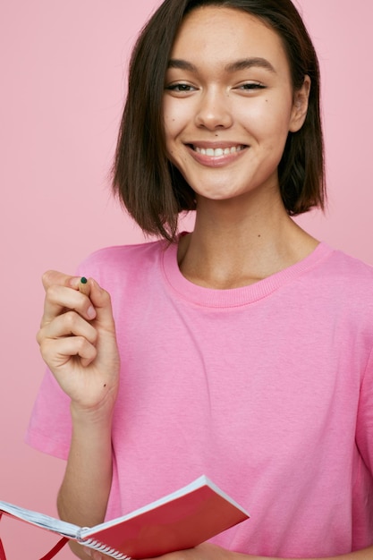 Short haired brunette in a pink tshirt red notebook and pen pink background