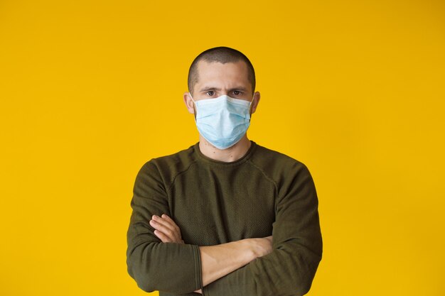 Short haired caucasian man wearing a white mask is posing on a yellow wall with crossed hands