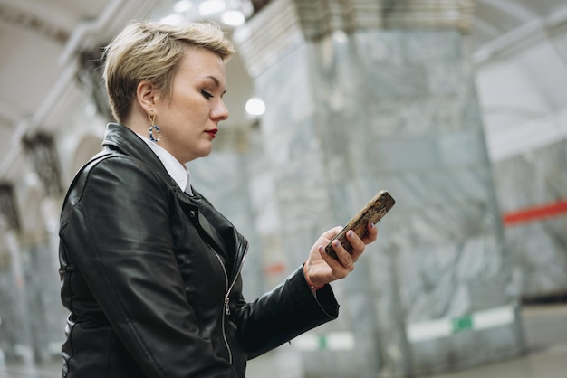 Short haired caucasian woman wearing leather jacket and jeans sitting alone on metro station reading something in smartphone waiting for train to come Image with selective focus and noise effect