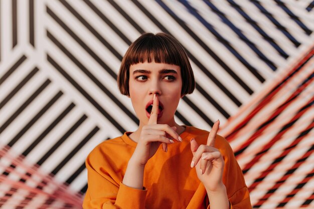 Short haired teen girl in bright clothes posing on striped background Modern woman in orange outfit looking away outside