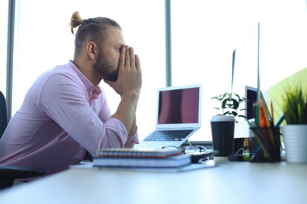 オフィスに座っている間、顔を手に保つスマートカジュアルウェアの疲れた若いビジネスマンの不足。