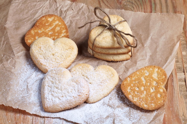 Shortbread cookies in shape of heart tied with a rope and several separate ones sprinkled with powdered sugar on paper located on a old wood