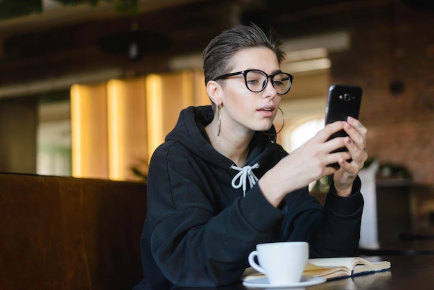 カフェテリアでコーヒーを飲みながらメッセージを入力するショートカットヒップスターの若いブロガー