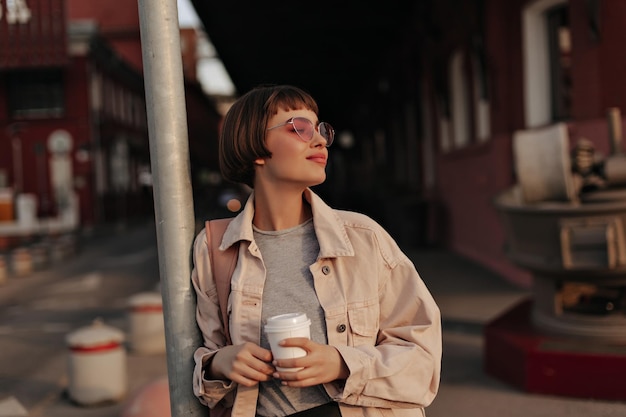 Photo shorthaired girl holding cup of tea outside trendy woman in sunglasses grey tshirt and light jacket posing at street