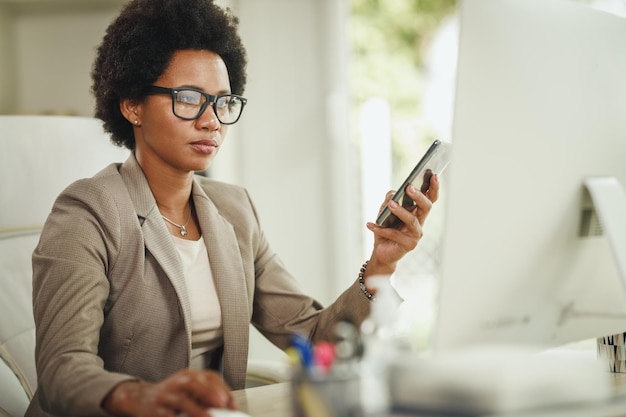 Shot of an African businesswoman using smartphone while working at computer in her home office during COVID-19 pandemic.