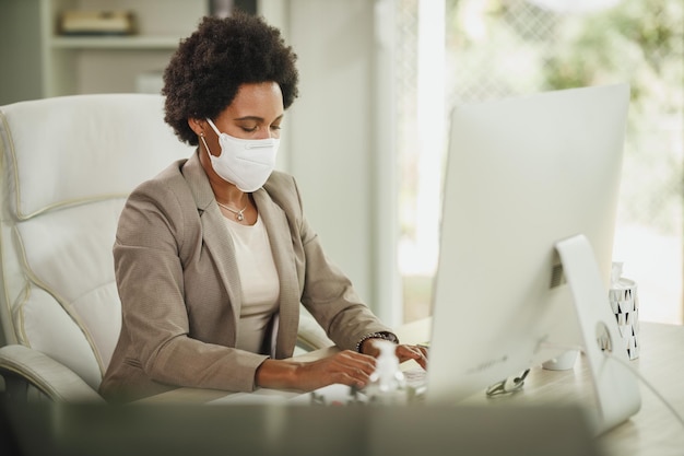 Photo shot of an african businesswoman with protective n95 mask sitting alone in her office and working on computer during corona virus pandemic.