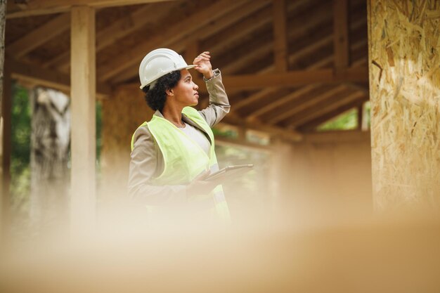 新しい木造住宅の建設現場をチェックしているアフリカの女性建築家のショット。彼女は保護用の作業服と白いヘルメットをかぶっています。