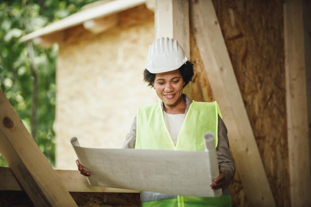 新しい木造住宅の建設現場で計画をチェックしているアフリカの女性建築家のショット。彼女は保護用の作業服と白いヘルメットをかぶっています。