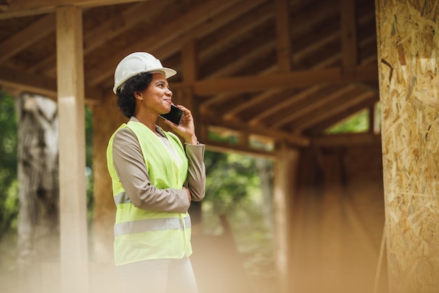 スマートフォンで話し、新しい木造住宅の建設現場をチェックしているアフリカの女性建築家のショット。彼女は保護用の作業服と白いヘルメットをかぶっています。