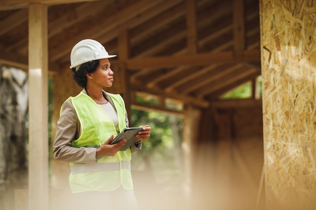 デジタルタブレットを使用し、新しい木造住宅の建設現場をチェックしているアフリカの女性建築家のショット。彼女は保護用の作業服と白いヘルメットをかぶっています。
