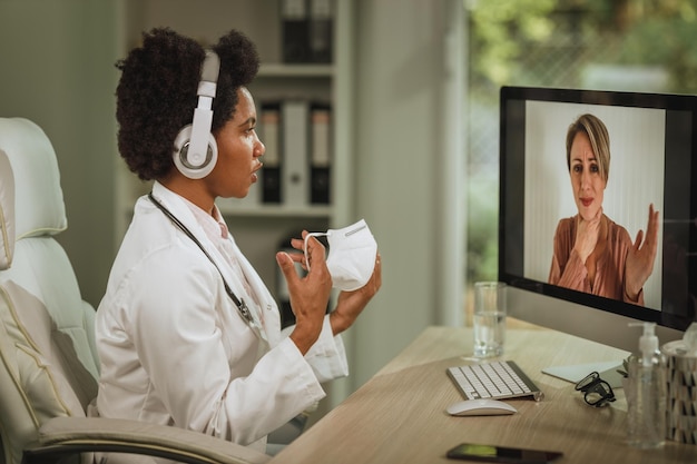 Photo shot of an african female doctor having video call with patient on computer in her consulting room during covid-19 pandemic.