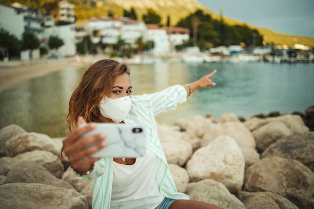 Shot of an attractive happy young woman wearing a protective\
n95 mask and making video call with her smartphone while spending\
time on the seaside during the covid-19.