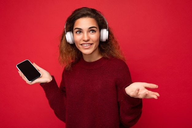 Shot of attractive positive young brunet curly woman wearing dark red sweater isolated on red