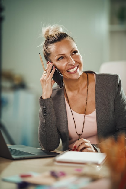 Shot of an attractive young businesswoman sitting alone in her home office and talking on smartphone during COVID-19 pandemic.