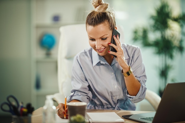 Photo shot of a attractive young businesswoman sitting alone in her home office and talking on smartphone during covid-19 pandemic.