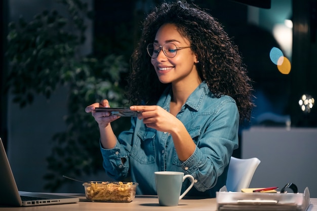 Shot of beautiful afro young business woman taking a photo with her smartphone while eating pasta sitting in the office.