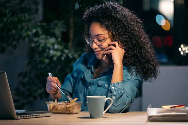 オフィスに座ってパスタを食べながらコンピューターで作業している美しいアフロ若いビジネス女性のショット。