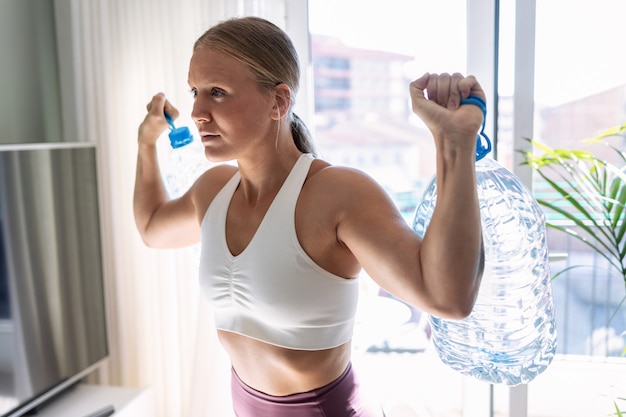 Shot of beautiful gymnast woman doing exercices lifting eight liters water bottles in living room at home.