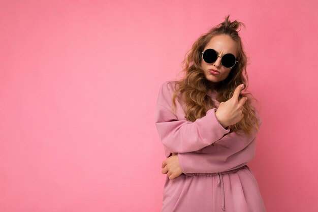 Shot of beautiful happy young dark blonde curly woman isolated over pink background wall wearing casual pink sport clothes and stylish sunglasses looking at camera and having fun. empty space