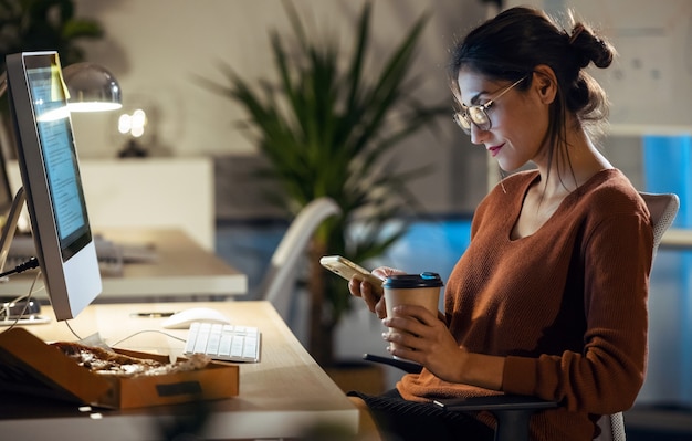 Shot of beautiful young business woman sending messages with mobile phone while working with computer sitting in the office.