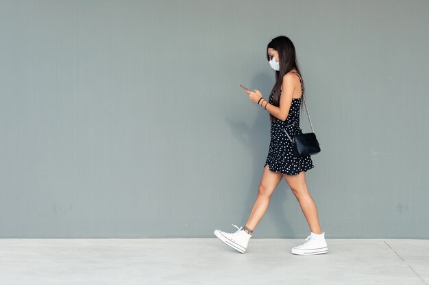 Shot of a beautiful young woman in face mask sending messages with her mobile phone while walking in the street.