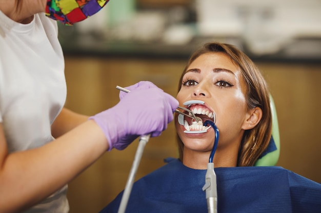 美しい若い女性のショットが歯科医にあります。彼女は歯科医の椅子に座り、歯科医は審美的な自己整列型舌側ロックを入れて歯にブレースをセットする準備をしています。