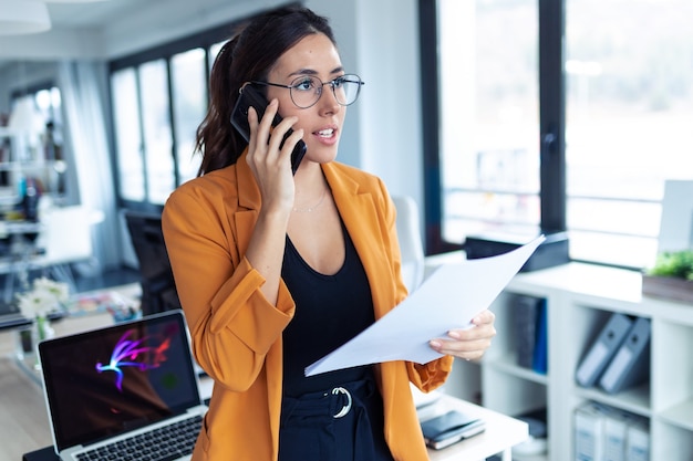 Shot of business young woman talking on mobile phone while holding papers in the office.