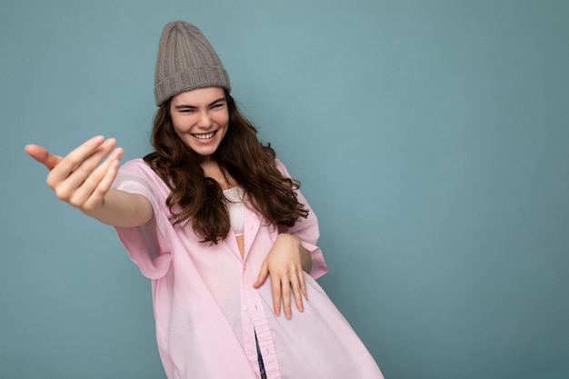 Shot of charming emotional young brunette female person standing isolated over blue background wall wearing pink shirt and gray hat looking at camera and having fun. Free space, copy space