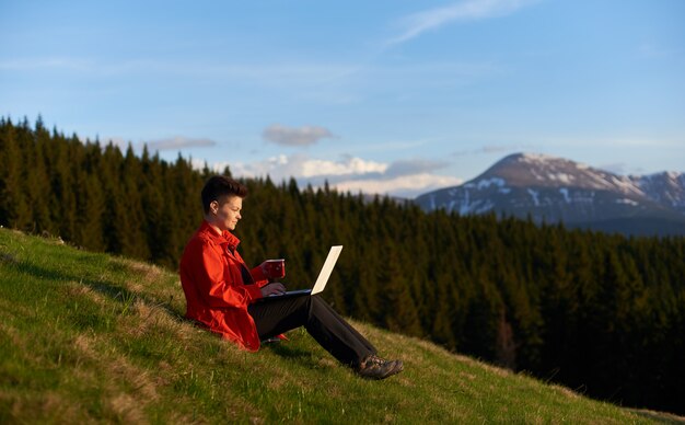 Снято жизнерадостной молодой женщины работая на ее компьтер-книжке отдыхая на верхней части горы на заходе солнца после пешего туризма