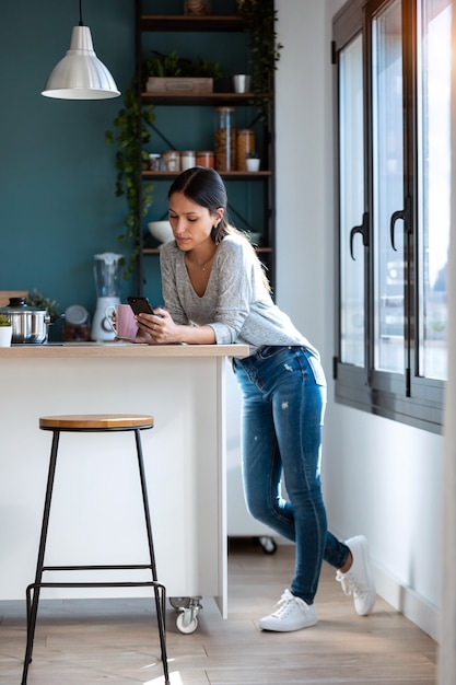 自宅のキッチンでコーヒーを飲みながら携帯電話を使用している集中した若い女性のショット。