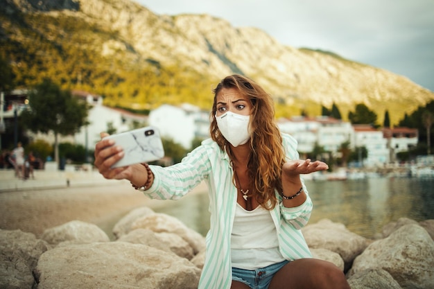 Shot of a concerned young woman wearing a protective n95 mask\
and making video call with her smartphone while spending time on\
the seaside during the covid-19.