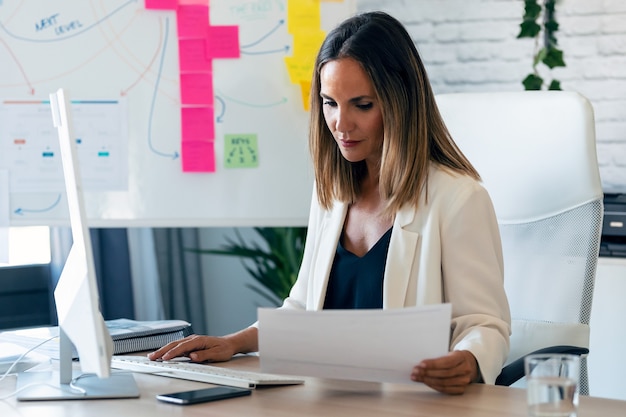 Shot of confident business woman working with computer while reading some notes in the office.