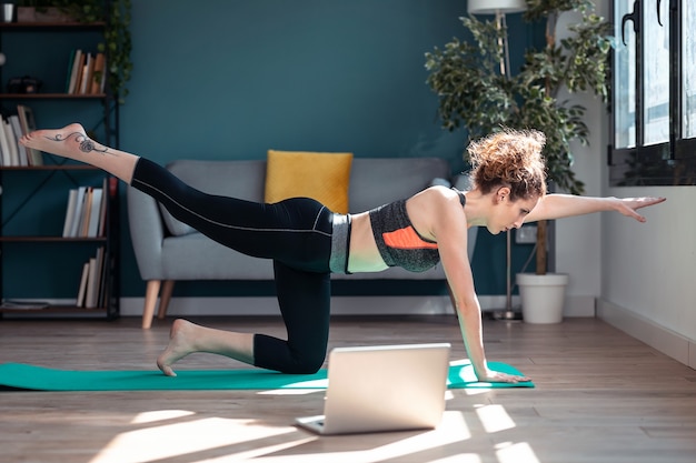 Shot of confident sporty young woman doing hypopressive exercises following online gym classes via laptop on floor in her living room at home.