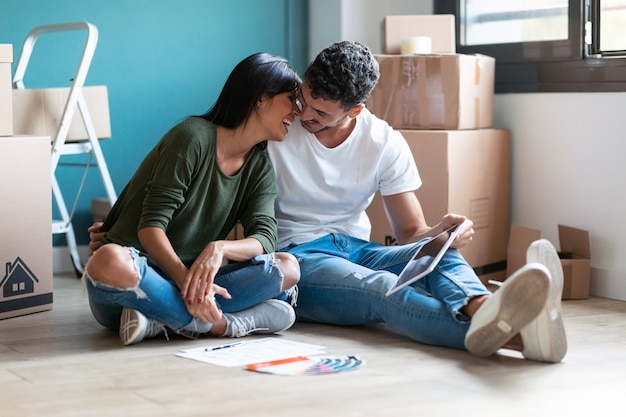 Снимок милой пары, целующейся друг с другом и ищущих идеи для украшения новой квартиры на цифровом планшете, сидящем на полу.