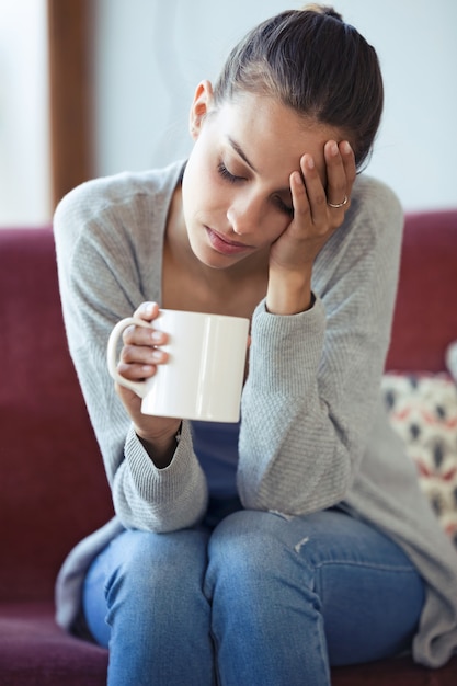 自宅のソファでコーヒーを飲みながら頭痛を抱えている落ち込んでいる若い女性のショット。