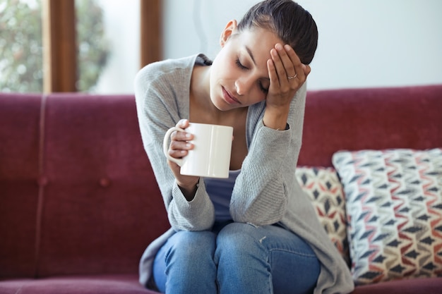 Shot of depressed young woman having headache while drinking coffee on sofa at home.