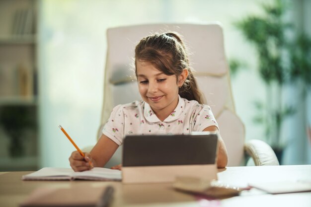 COVID-19 전염병 동안 집에서 온라인 수업을 하기 위해 태블릿을 사용하는 부지런한 어린 소녀의 사진.