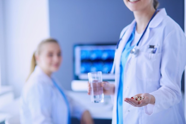 Снимок врача, держащего таблетки и стакан воды в клинике с коллегой на заднем плане.