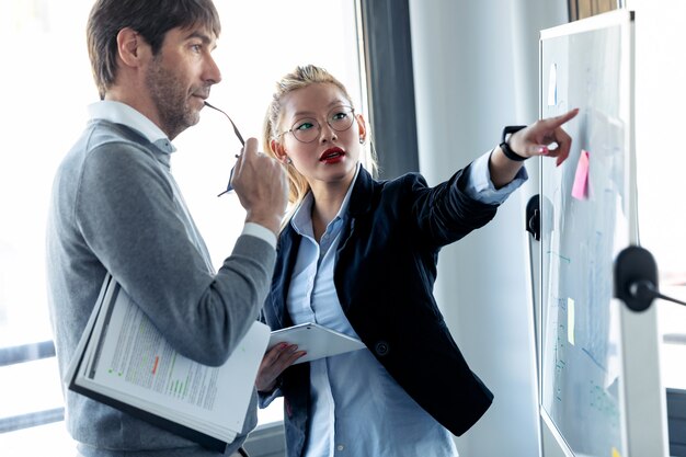 Shot of elegant young businesswoman pointing at whiteboard and explaining to the project to her colleague on coworking place.