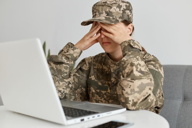 Shot of exhausted woman soldier wearing camouflage uniform and\
hat, posing at home, sitting at table with laptop on it, covering\
eyes with palms, having eyes hurt and headache, long hours\
working.