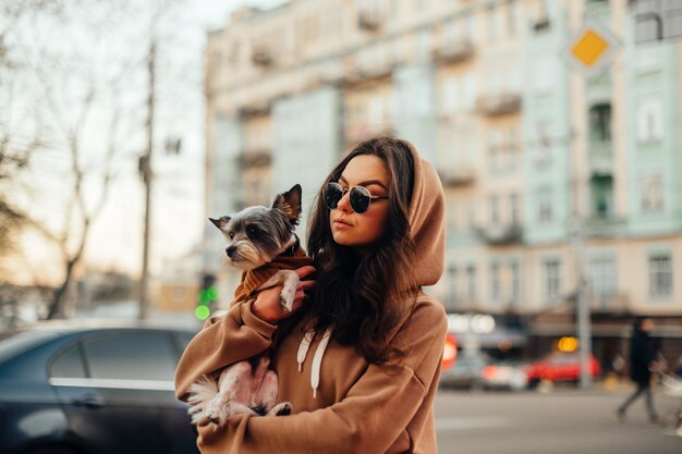 Inquadratura di una donna alla moda in occhiali da sole e felpa con cappuccio elegante che abbraccia il suo piccolo cane york terrier